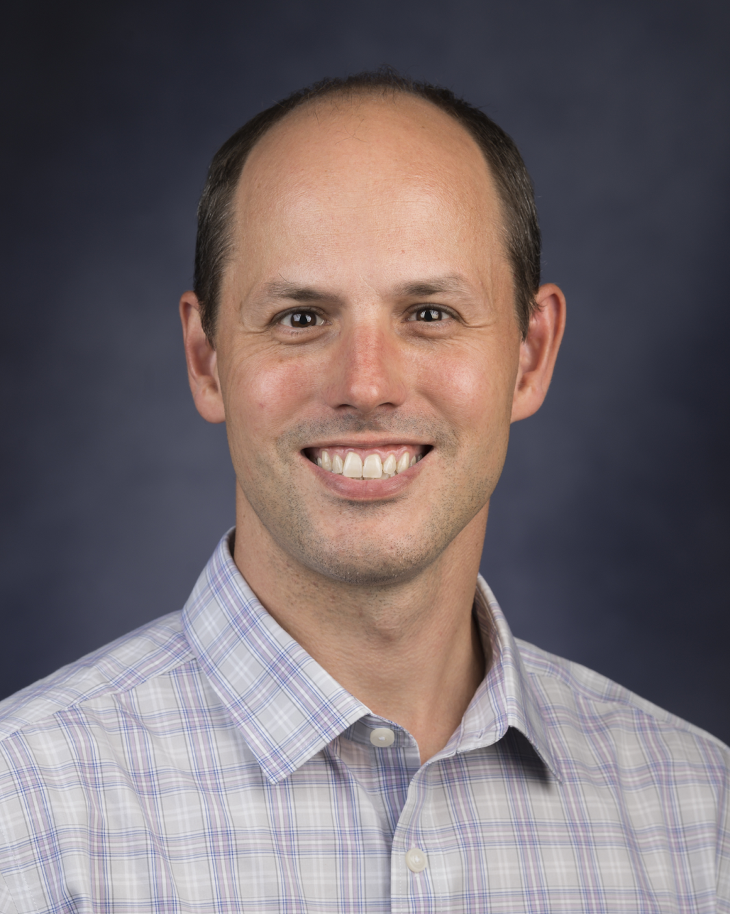 A smiling White man in a white and blue checked button-down shirt.